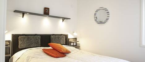 Una cama bien hecha con un edredón blanco y dos almohadas naranjas, un cabecero de madera y una estantería de pared con un cuadro.