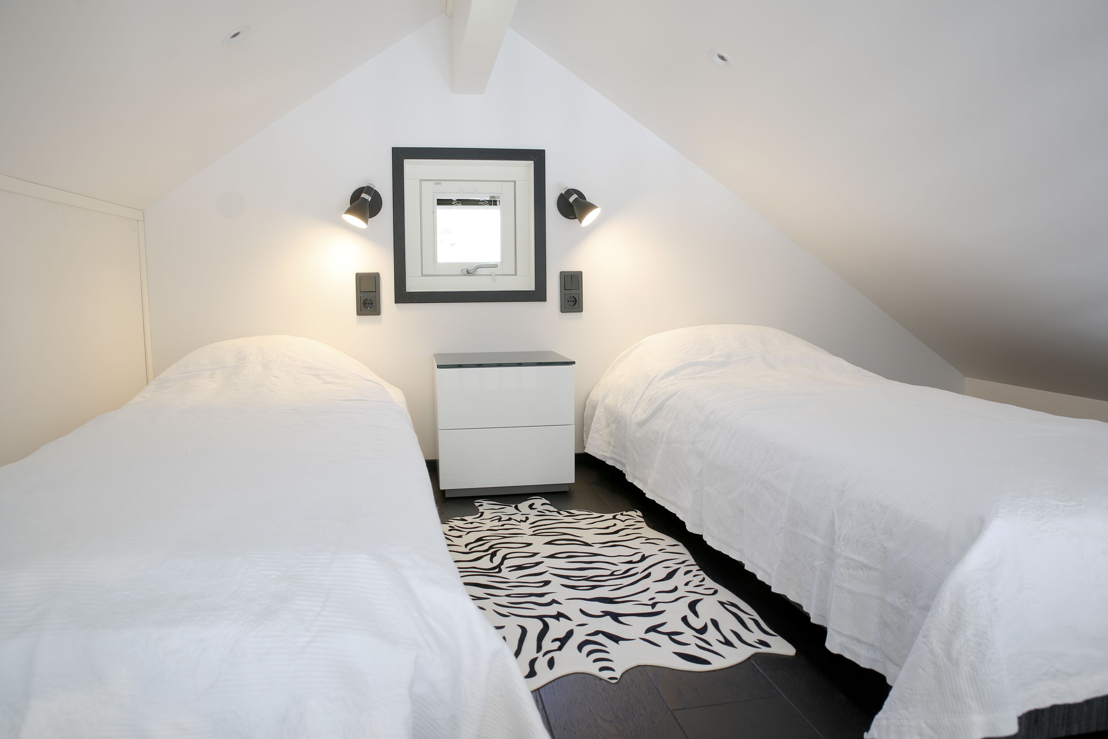 A small, triangular room with two single beds, a nightstand, a mirror, and a zebra-patterned rug.