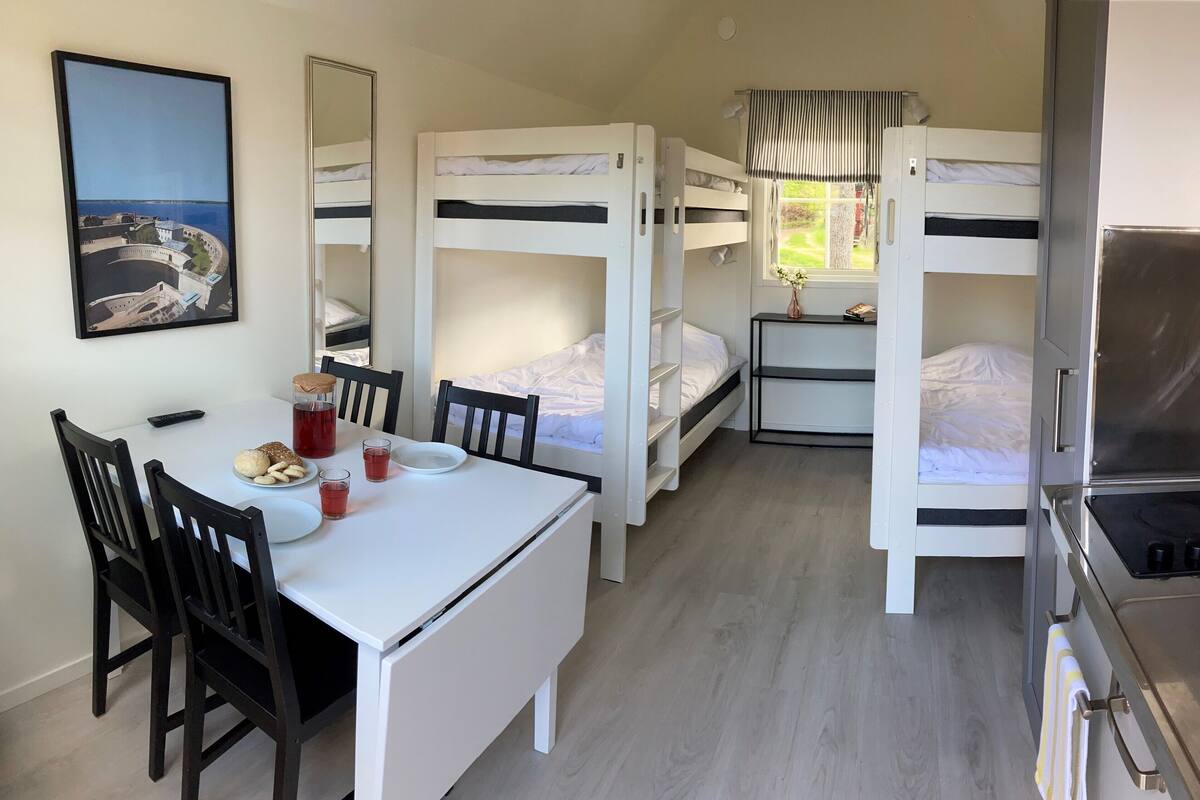A compact room with bunk beds, a small dining table, chairs, a kitchen area with a stove, and a framed picture on the wall.