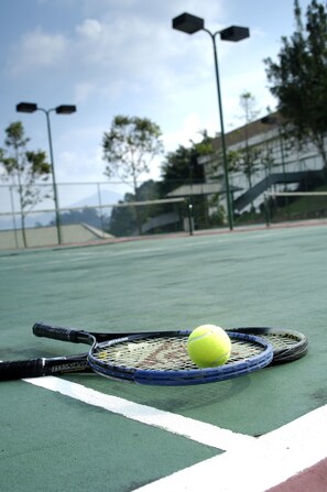 A tennis court with a tennis racket and ball, a fence, and trees in the background.