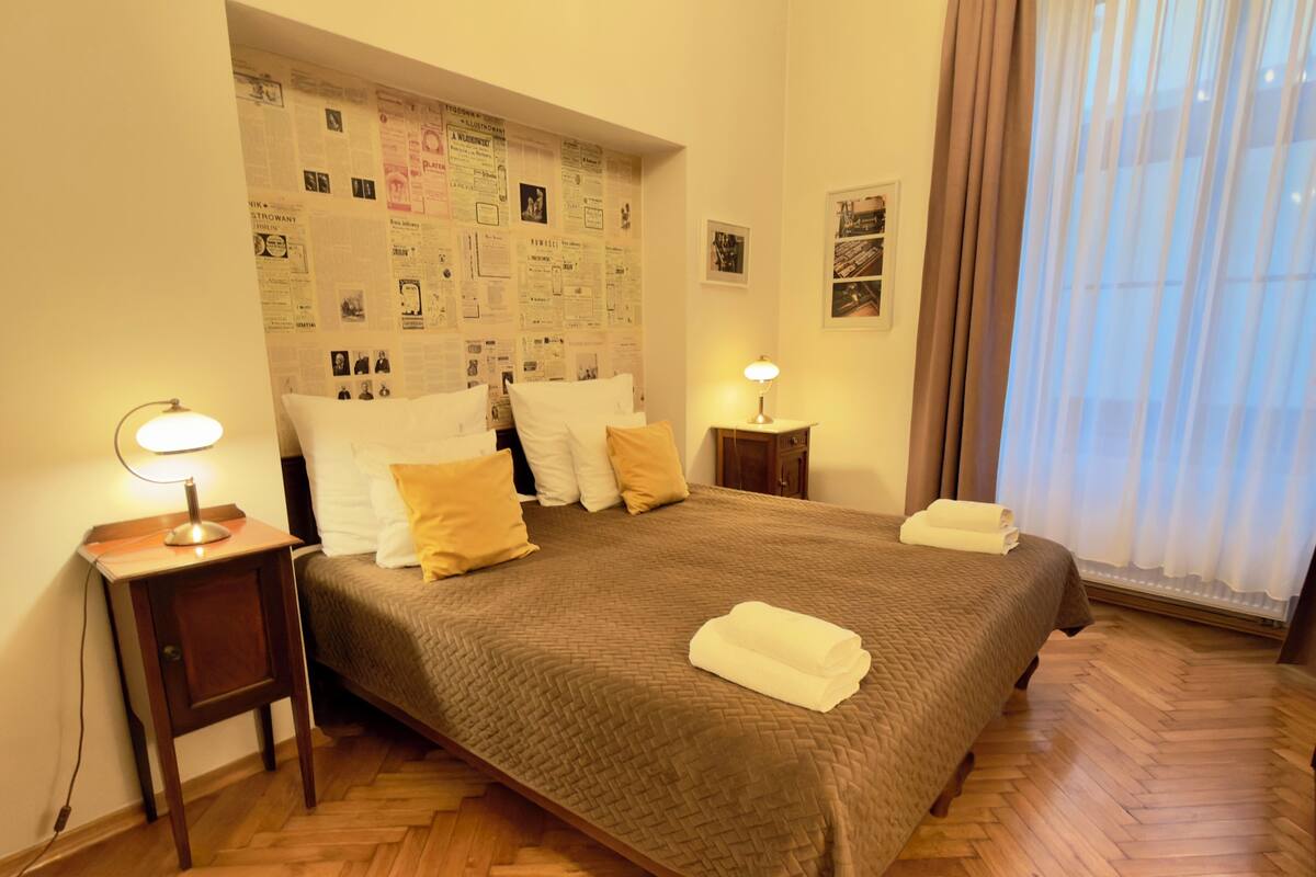 A hotel room with a large bed, a headboard with posters, a bedside table with a lamp, and a window with curtains.