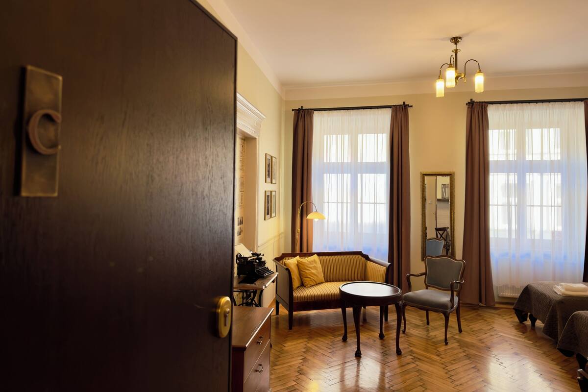 A room with a wooden floor, a sofa, a chair, a small table, and a bed.