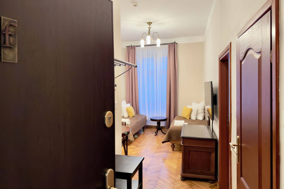 A hotel room with a wooden floor, a bed with pillows, a small table, a chair, a TV, and a window with curtains.