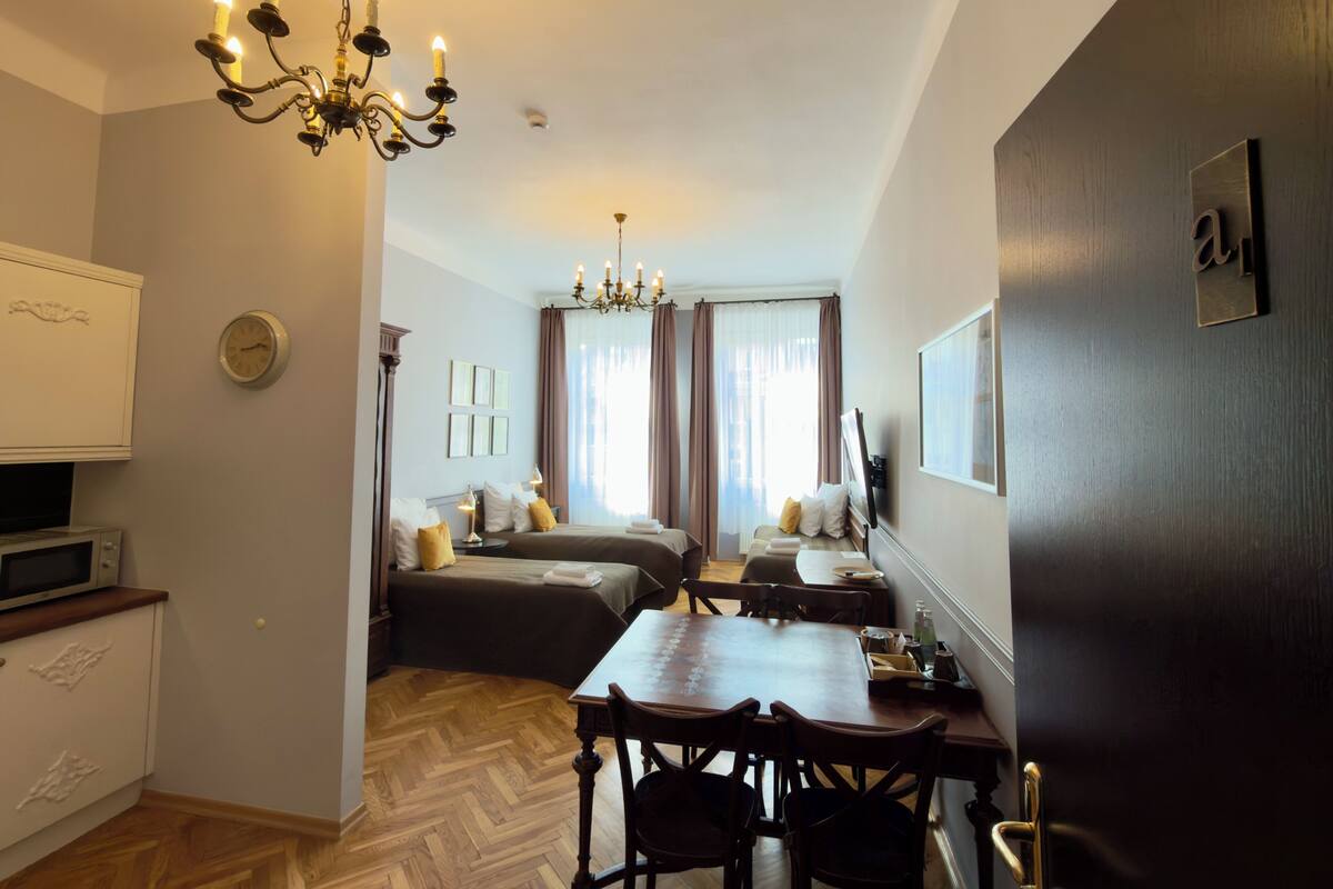 A hotel room with a dining area, a chandelier, a clock, and a bed with pillows.
