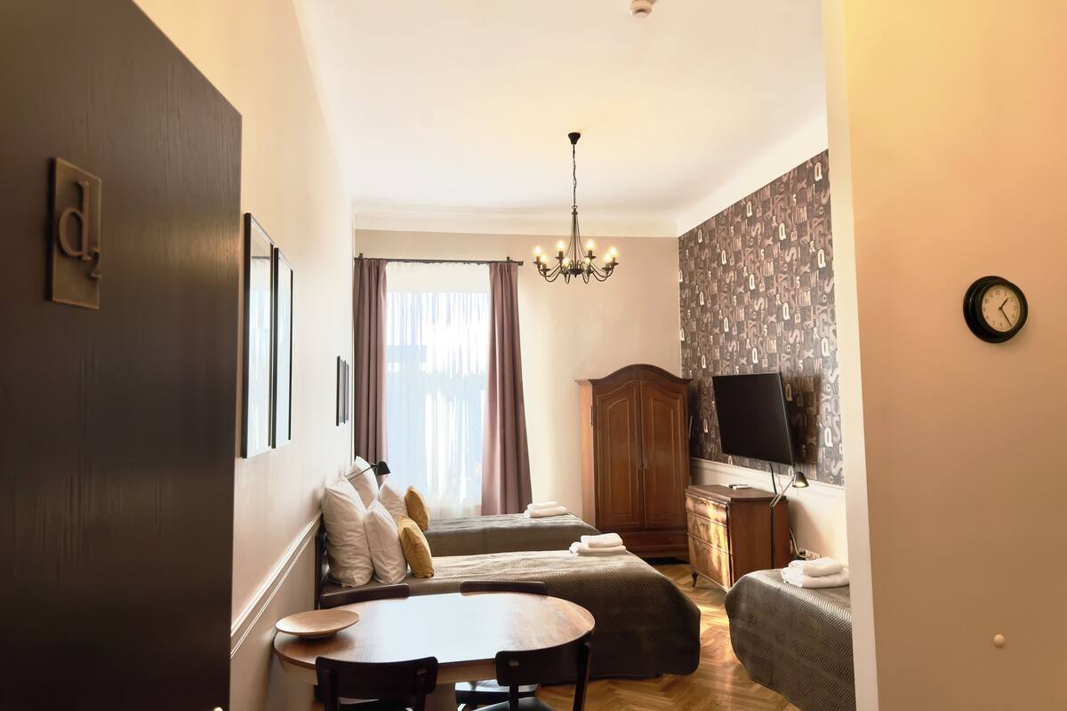 A compact hotel room with a bed, a small dining table, a TV, and a chandelier.