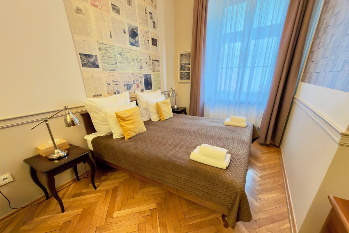 A neatly arranged bedroom with a bed, bedside table, lamp, and a wall adorned with various papers.