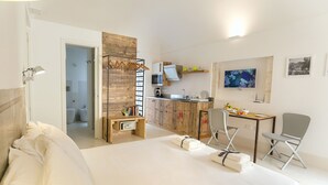 A hotel room with a bed, a desk, a chair, a kitchenette, and a bathroom visible through an open door. - Dione Apartments Ortigia (Syracuse)