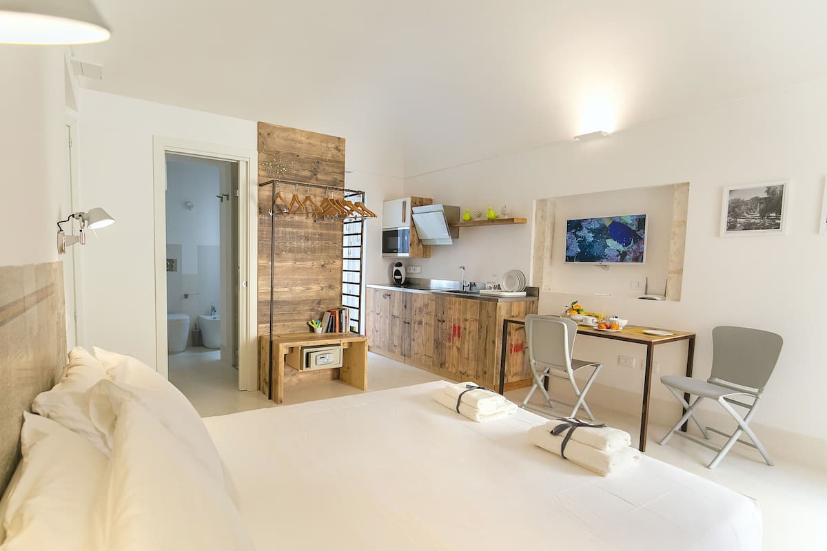 Quarto de hotel com cama, escrivaninha, cadeira, cozinha compacta e banheiro visível por uma porta aberta.