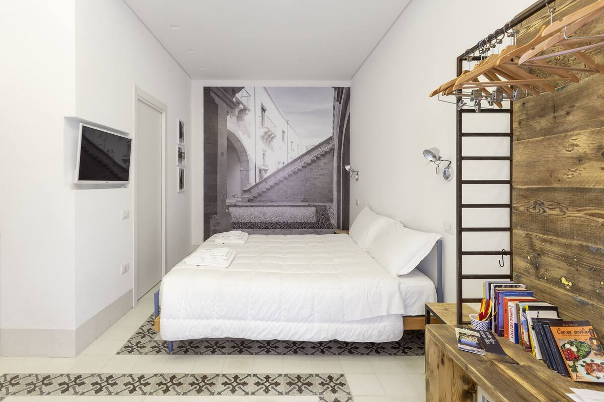 Quarto com uma cama, uma escada de madeira, uma televisão e livros sobre uma mesa lateral.