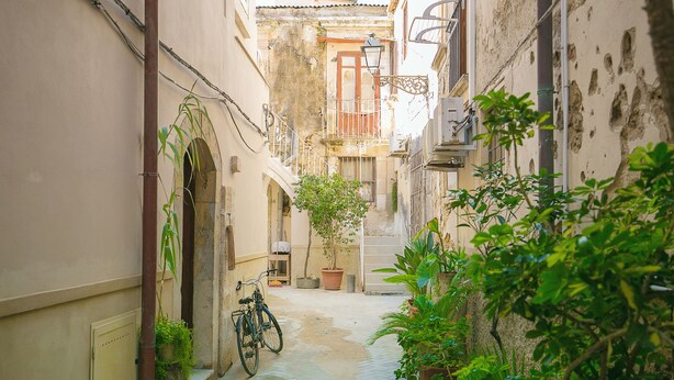 A narrow cobblestone alley with a bicycle parked on the left, surrounded by lush greenery and traditional architecture. - Dione Apartments Ortigia (Syracuse)