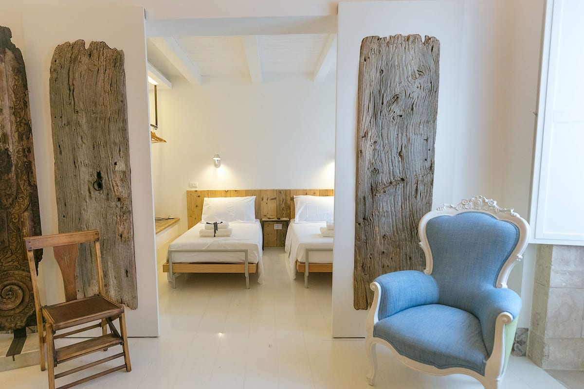 Quarto de hotel com cabeceira e cadeira de madeira, e duas camas com lençóis brancos.