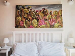 A bedroom with a white bed, white nightstands, and a painting of cacti and flowers on the wall. - Family Apartments (Syracuse)