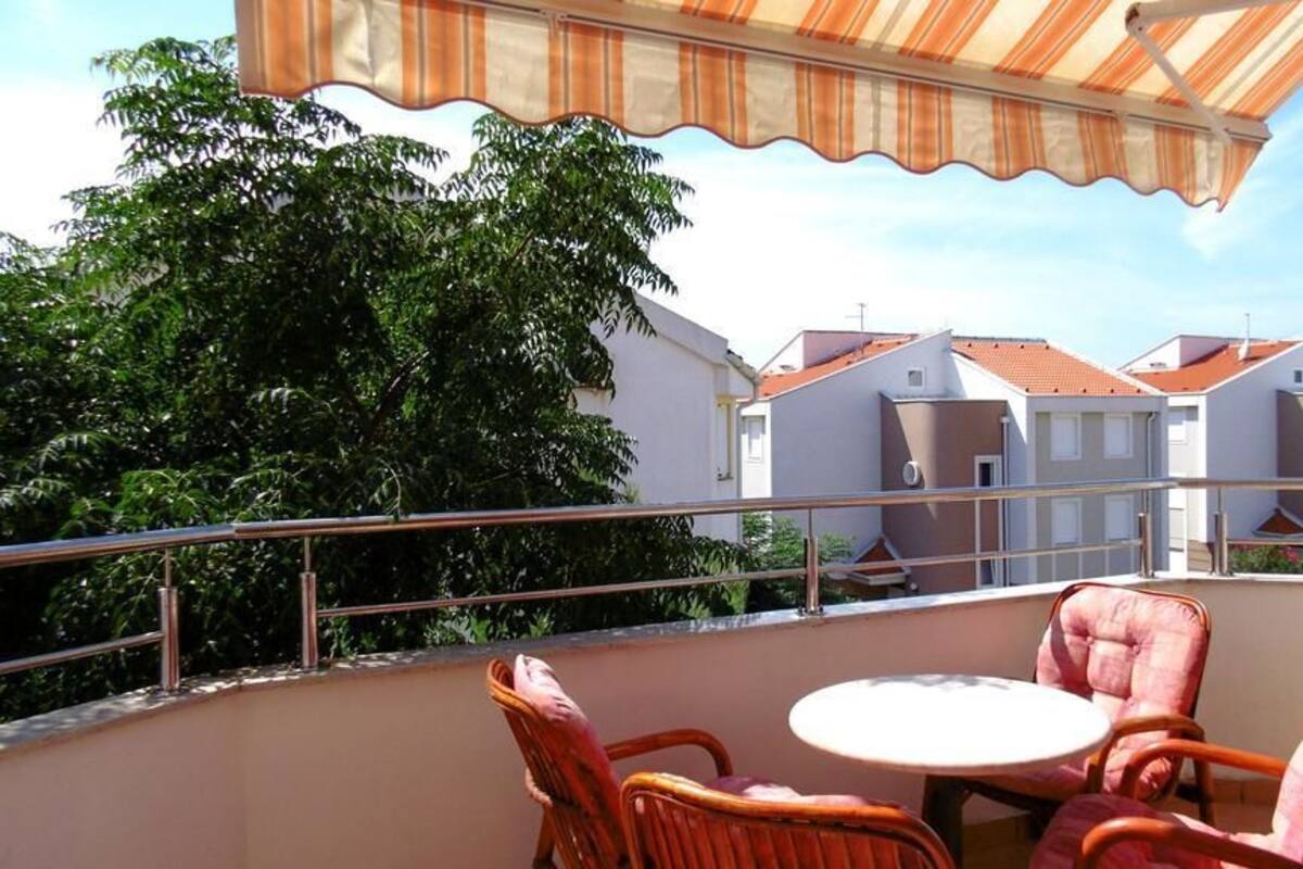 A balcony with wicker chairs and a table, overlooking a row of houses with red roofs.