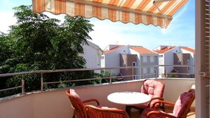 A balcony with wicker chairs and a table, overlooking a row of houses with red roofs.