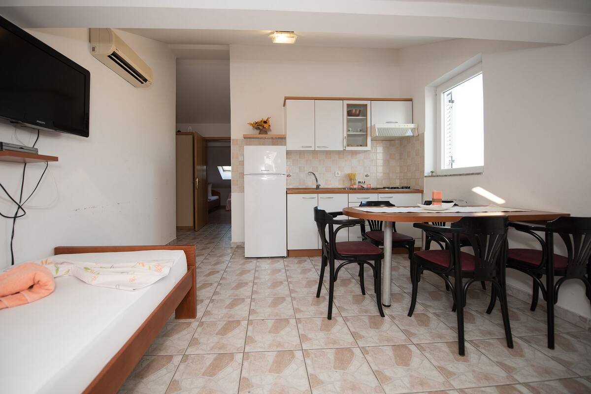 A compact kitchen with a dining area, a TV mounted on the wall, and a bed with a pillow.