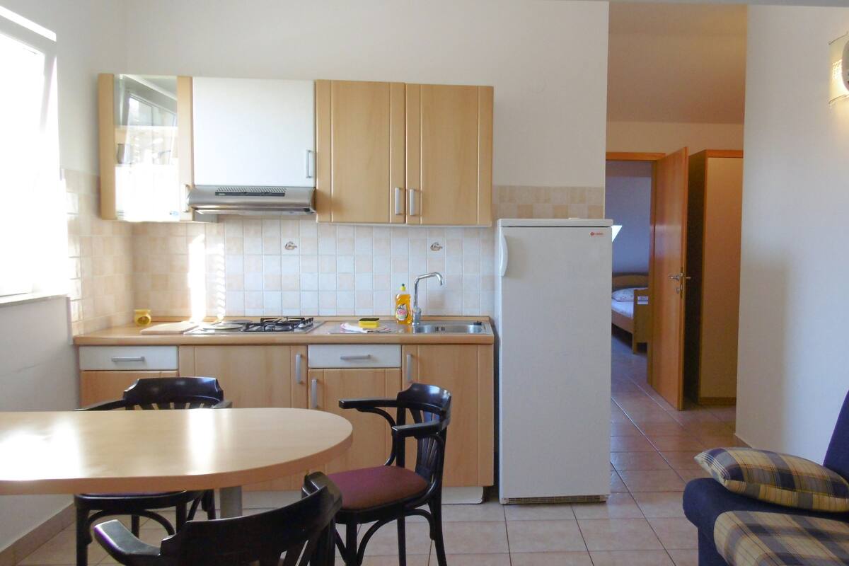 A compact kitchen with wooden cabinets, a white refrigerator, a dining table with chairs, and a sofa in the adjacent living area.
