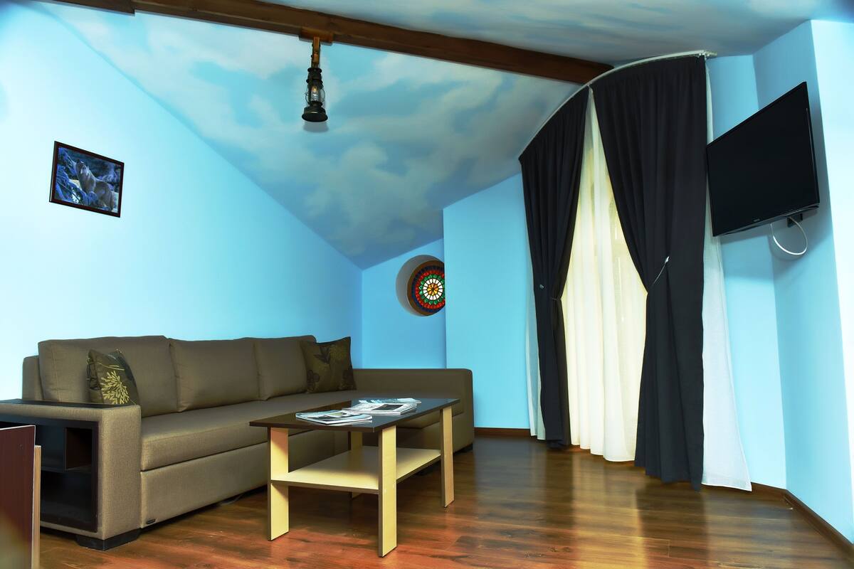 A living room with a sofa, a coffee table, a flat-screen TV mounted on the wall, a ceiling fan, and a window with curtains.