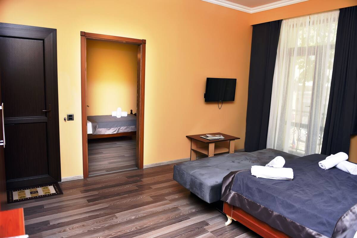 A hotel room with a wooden floor, a bed with a dark blue bedspread, a second bed, a small table with a remote control, and a television on the wall.