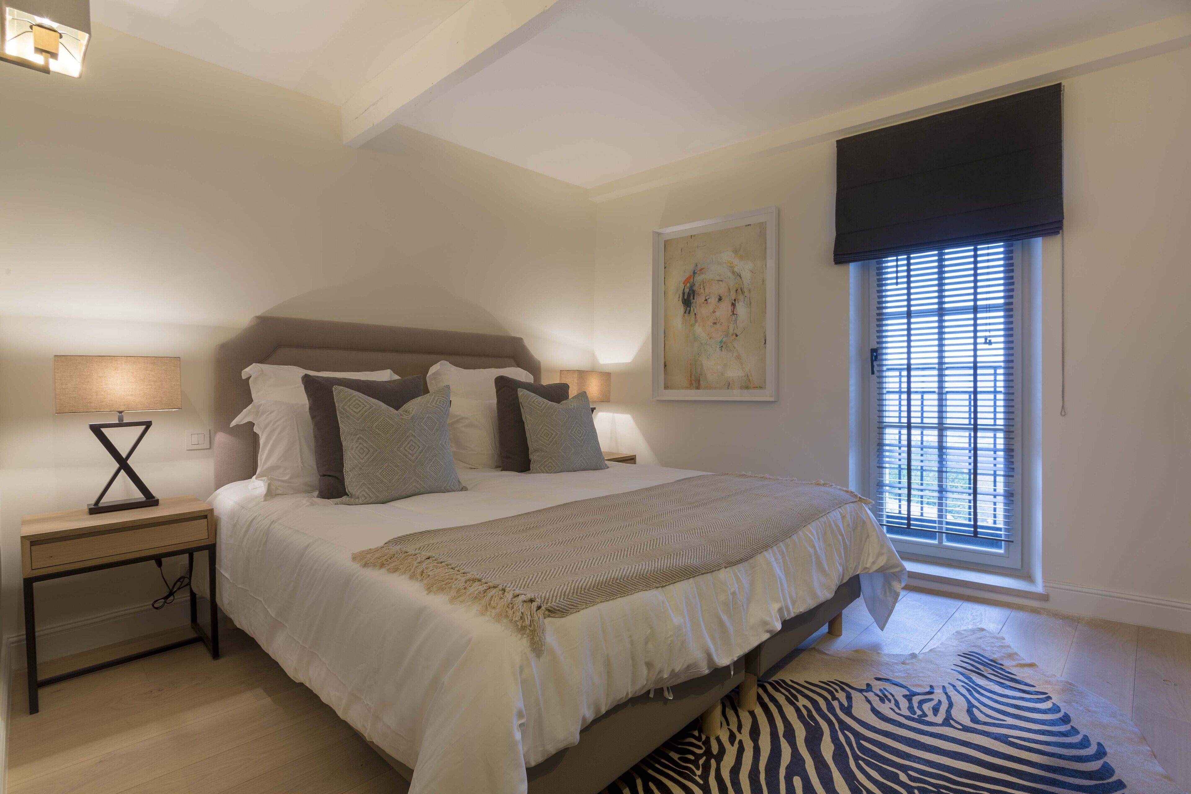 A bedroom with a bed, bedside tables, a lamp, a window with blinds, and a zebra-patterned rug.