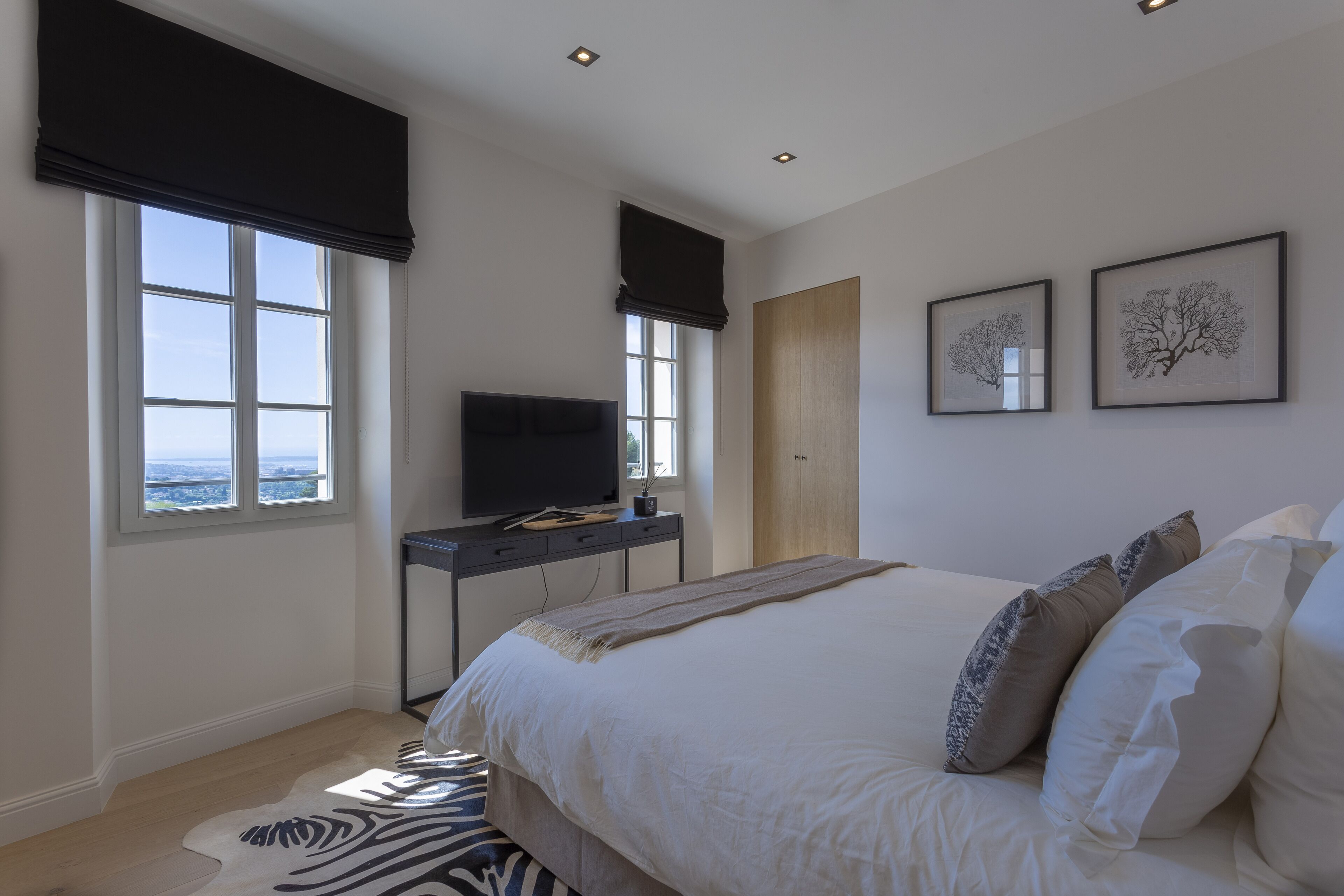 A bedroom with a large bed, a desk with a TV, two framed pictures on the wall, and a window with a view.