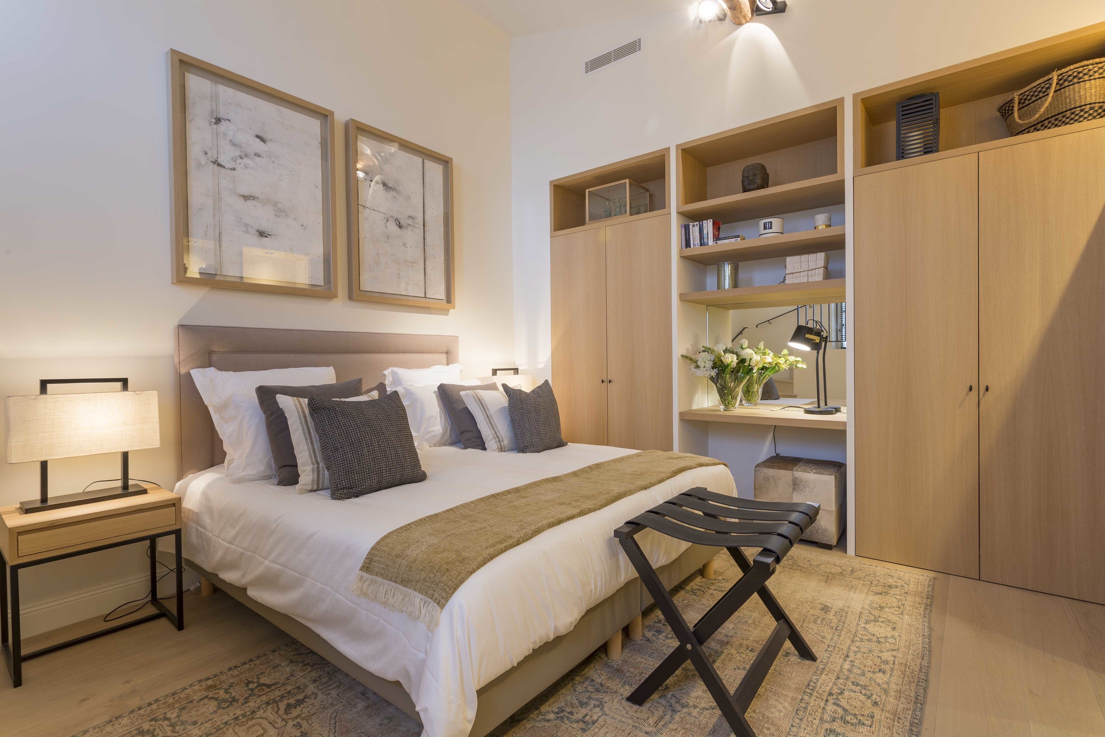 A bedroom with a bed, bedside table, lamp, and a bench. There are wooden cabinets, a shelf with books, and a vase with flowers.