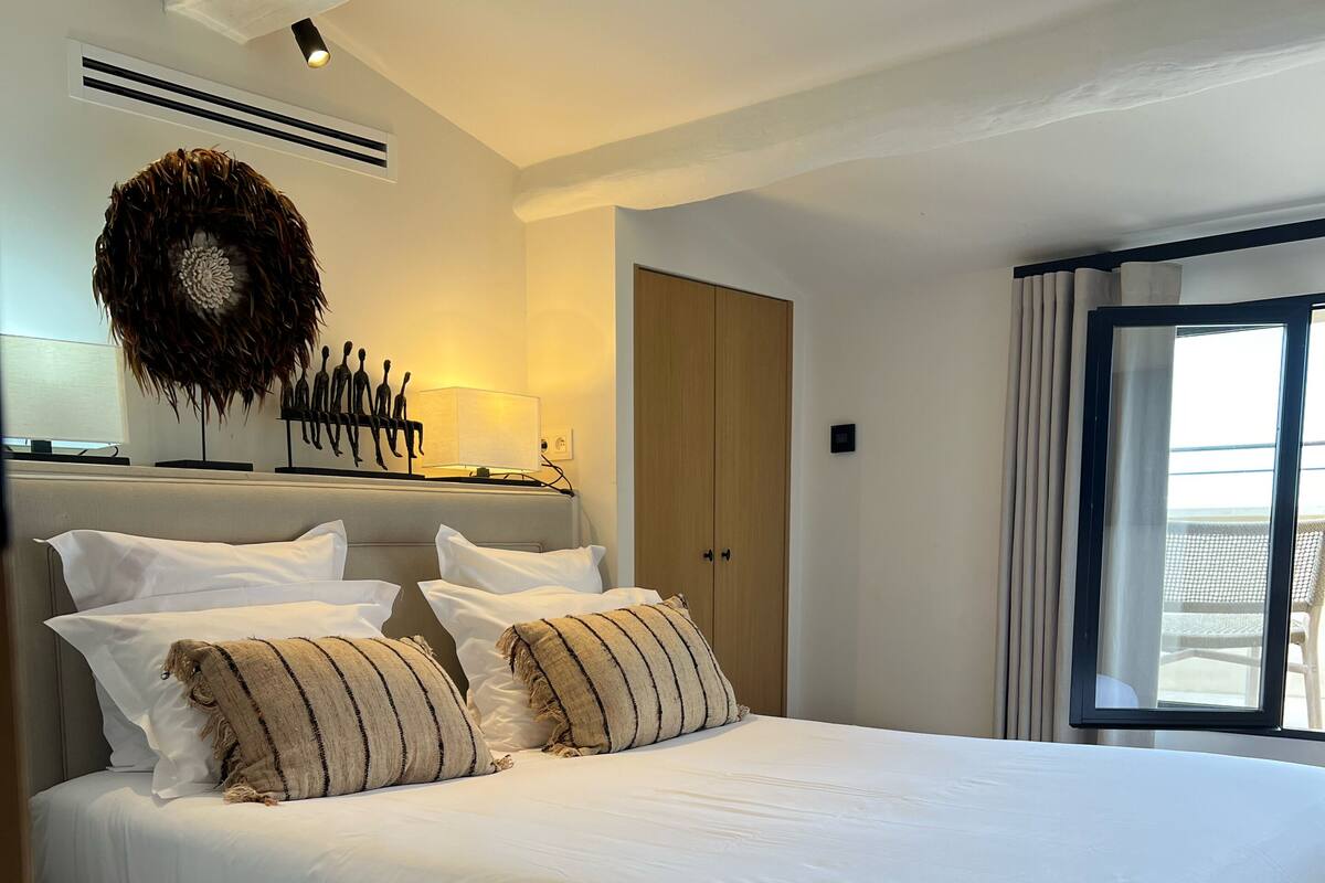 Uma cama grande com lençóis brancos, cabeceira com detalhe decorativo na parede e guarda-roupa de madeira.