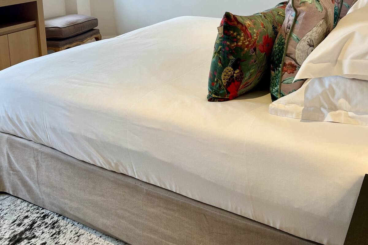 Cama bem arrumada com lençóis brancos, cabeceira de madeira e almofadas estampadas com flores. Há duas obras de arte emolduradas na parede, uma janela com cortina azul escura e uma mesa de cabeceira de madeira com abajur.