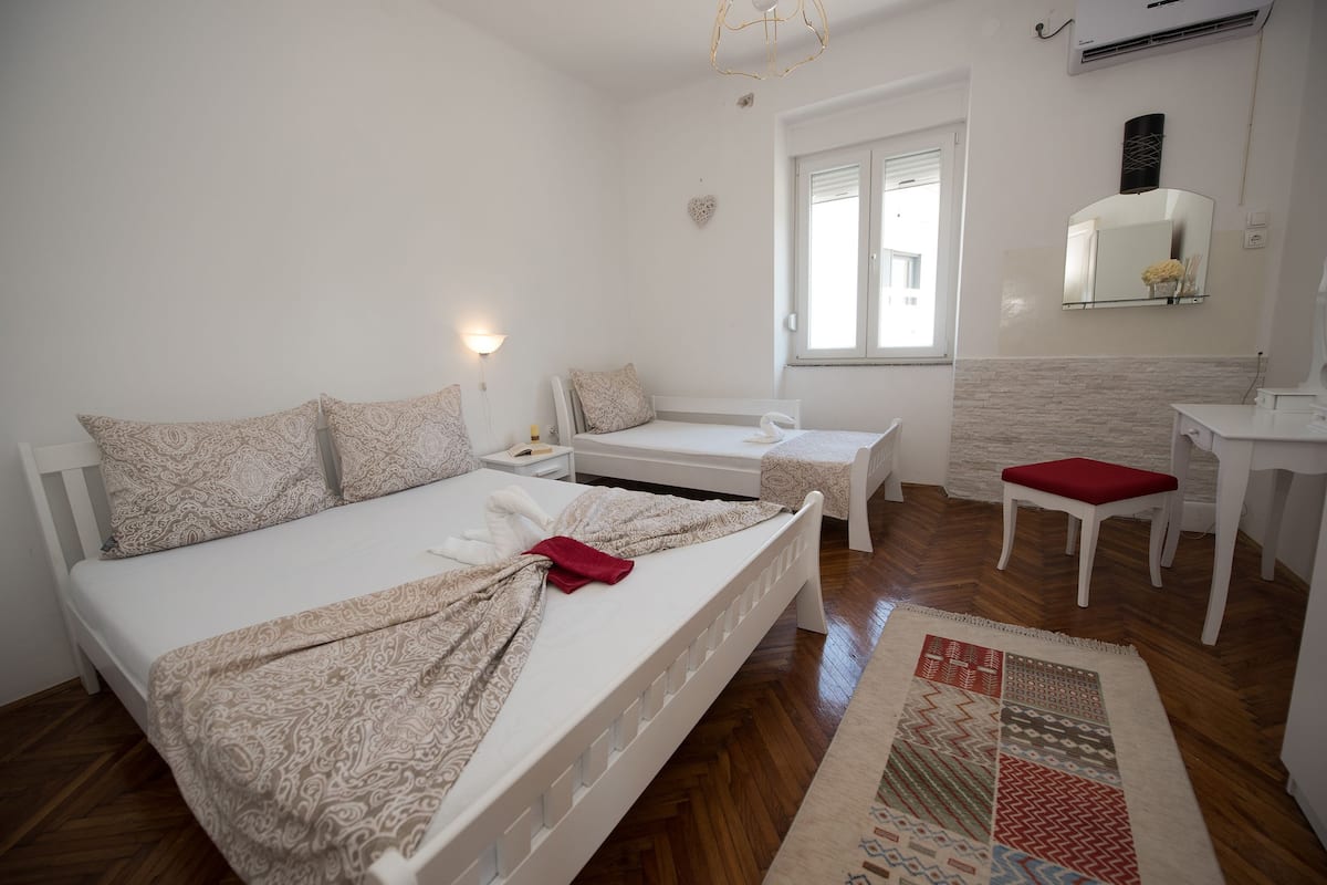 Ein helles Schlafzimmer mit einem großen Bett, einem Einzelbett, einem Schreibtisch und einem Stuhl. Ein Fenster lässt natürliches Licht herein und über dem Schreibtisch befindet sich ein Spiegel.
