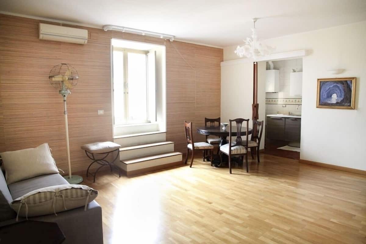 Amplia sala de estar con piso de madera, un sofá, una mesa de comedor con sillas, una escalera y una cocina visible a través de una puerta abierta.