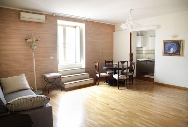 A spacious living area with a wooden floor, a sofa, a dining table with chairs, a staircase, and a kitchen visible through an open door.