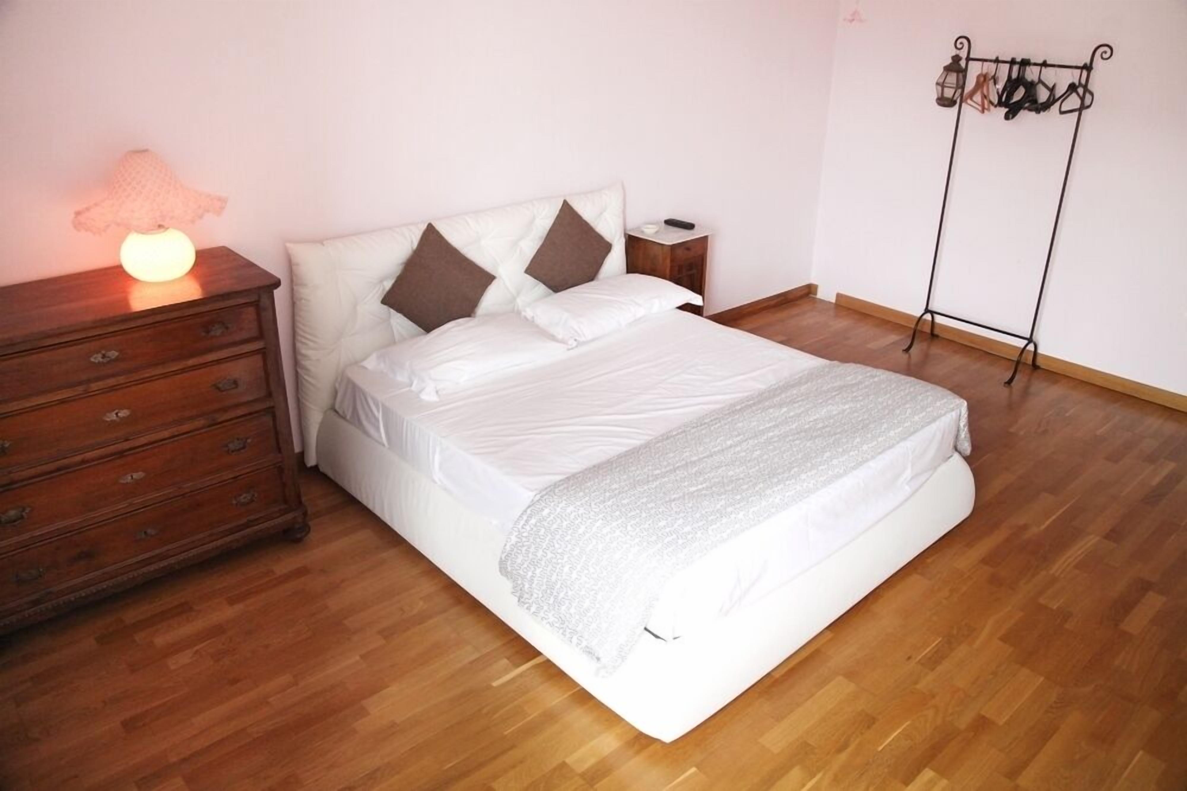 A neatly made bed with a white comforter and pillows, a wooden dresser with drawers, a bedside table with a lamp, and a coat rack against the wall.