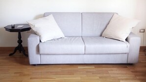 A light grey sofa with two white cushions, a small black side table with a remote control, and a wooden floor. - Gramsci Apartments (Naples)