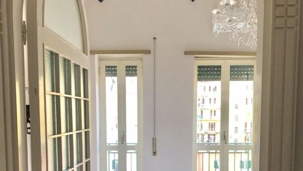 A hotel room with a bed, a chandelier, and a view of buildings through the windows. - Gramsci Apartments (Naples)