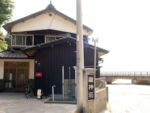 Un edificio de estilo arquitectónico japonés tradicional, con tejado de tejas y fachada de madera. Hay una bicicleta estacionada afuera, y un letrero con caracteres japoneses es visible en el lado derecho del edificio.