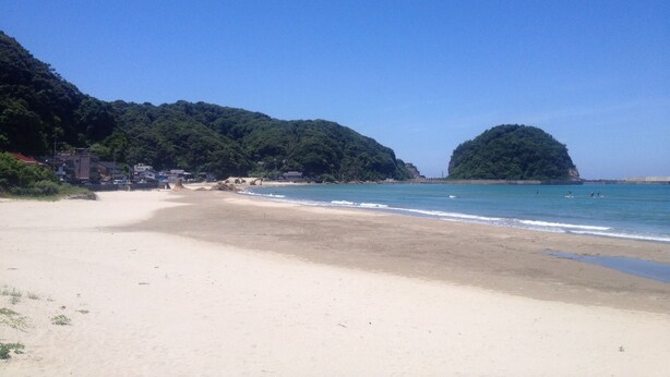 Una playa con agua azul cristalina, orilla de arena y colinas verdes exuberantes al fondo.