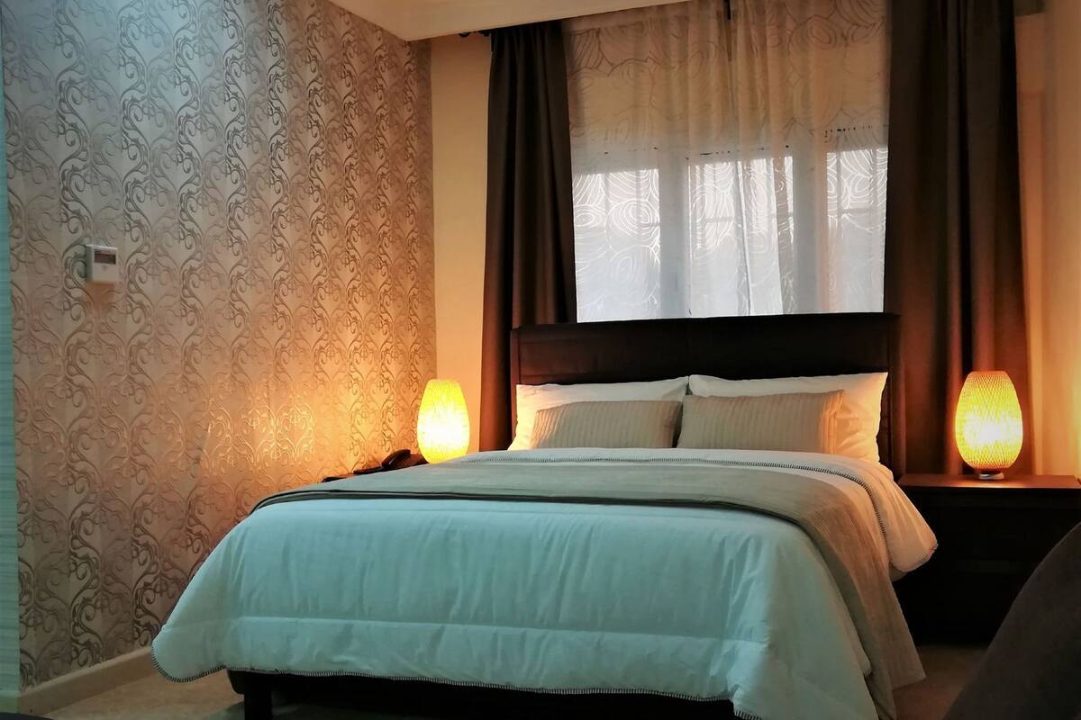 A neatly made bed with a headboard, two bedside lamps, and patterned wallpaper.