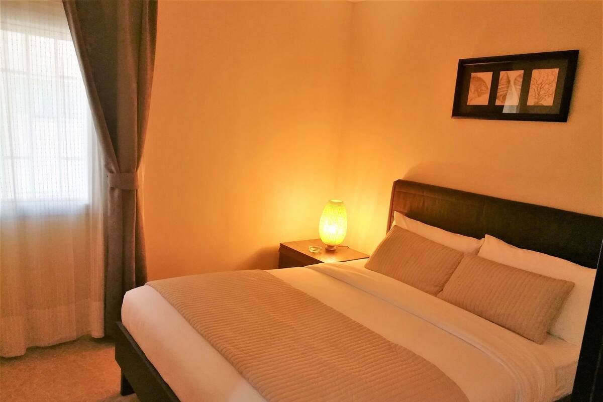 A hotel room with a bed, a nightstand with a lamp, a framed picture on the wall, and a window with curtains.