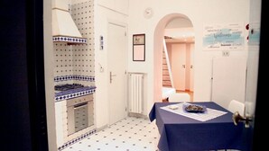 A compact kitchen with a stove, oven, and tiled backsplash. A dining table with a blue tablecloth and a bowl of fruit is visible. - Luxury House (Naples)