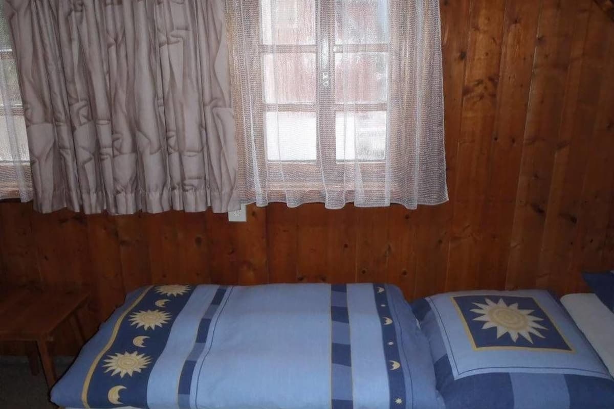 Quarto com paredes de madeira, cama com roupa de cama azul com motivos de sol e lua, e janela com cortina branca.
