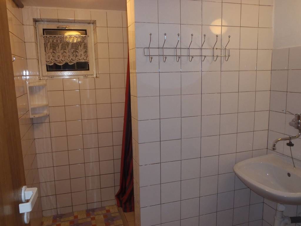 A bathroom with white tiled walls, a window with lace curtains, a towel rack, a sink, and a shower area.