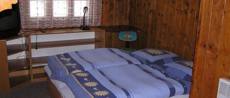 Un dormitorio con pared de madera, cama con ropa de cama azul y blanca, escritorio con lámpara y ventana con cortinas de encaje.