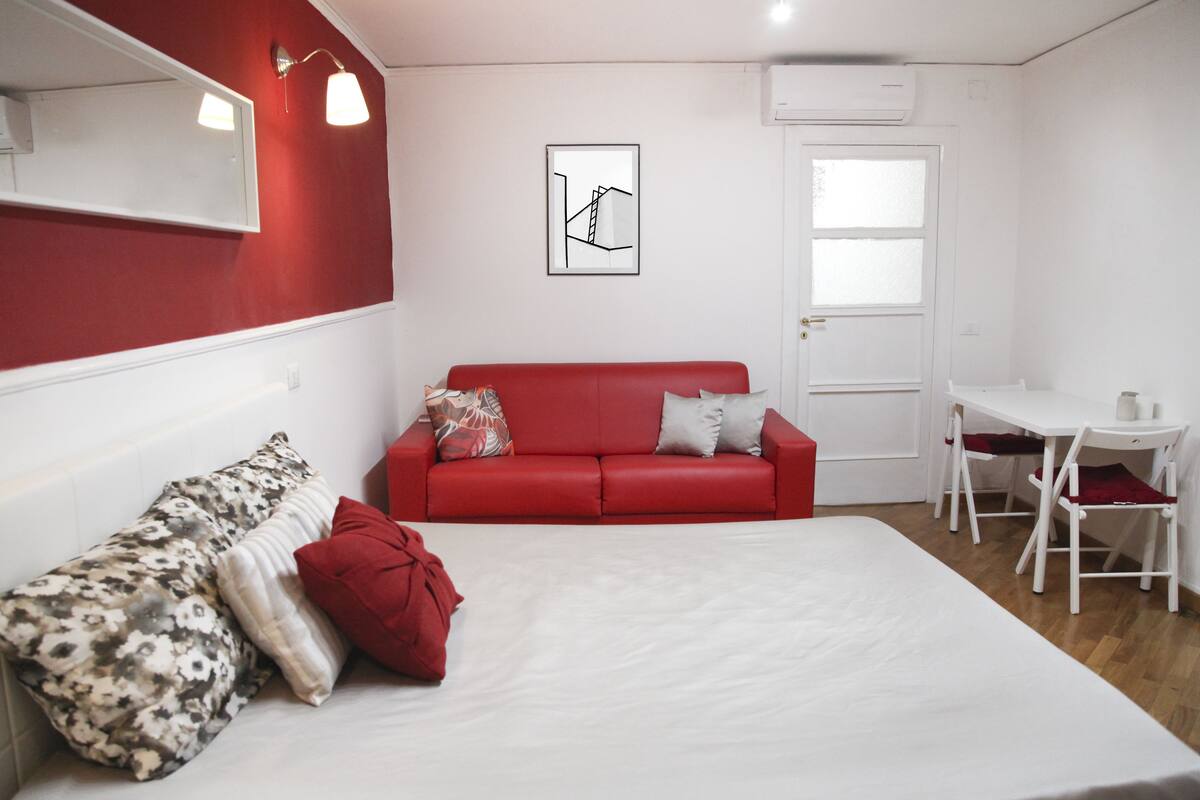 A room with a bed, a red sofa, a small white table, and a chair.