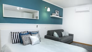 A bedroom with a bed, a gray sofa, a white wall, a mirror, and a shelf with items. - Colorful Sea View Apartments (Naples)