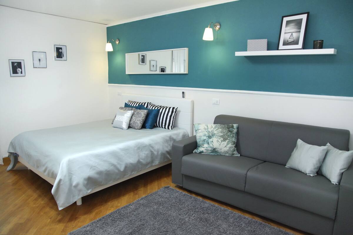 A bedroom with a bed, a gray sofa, a teal accent wall, a white shelf with items, and a mirror.