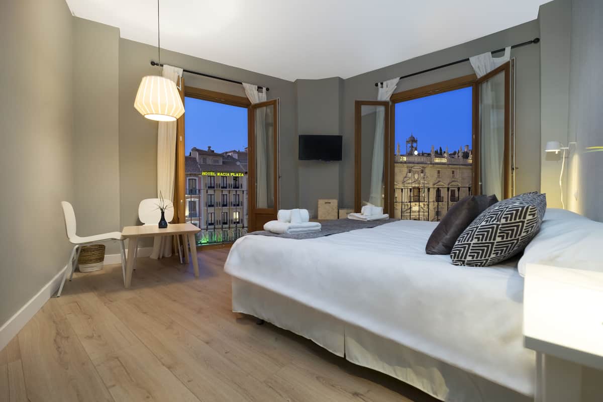 Ein Hotelzimmer mit einem großen Bett, einem Schreibtisch und einem Stuhl. An der Wand ist ein Fernseher befestigt und es gibt einen Balkon mit Blick auf Gebäude.