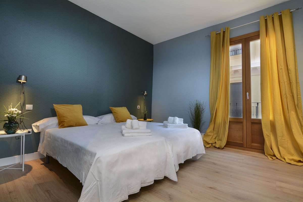 Ein Hotelzimmer mit zwei Betten, einem kleinen Tisch mit einer Vase und einem Fenster mit gelben Vorhängen.