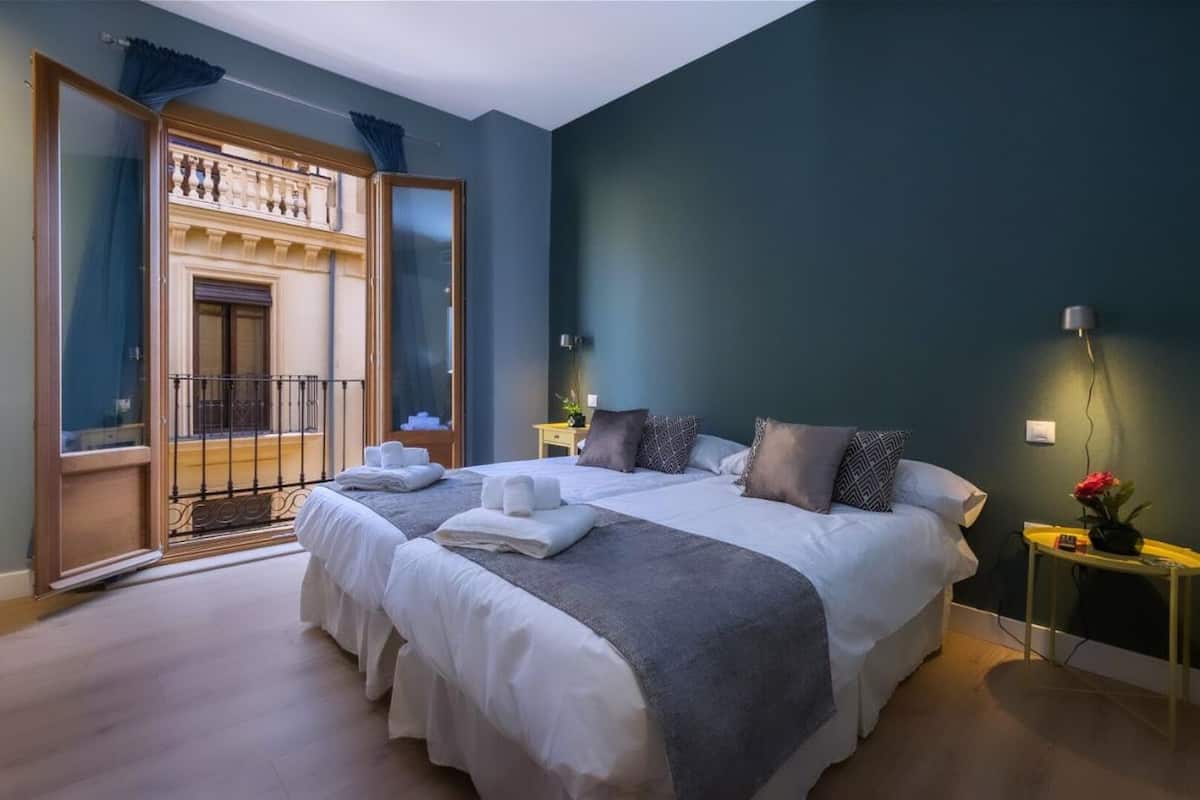 Ein Doppelbett mit weißen Leinentüchern und einer grauen Decke, ein Nachttisch mit einer Lampe sowie der Blick auf einen Balkon mit Geländer.