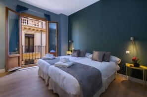 Un lit double avec du linge de lit blanc et un plaid gris, une table de chevet avec une lampe, et une vue sur un balcon doté d’une balustrade.
