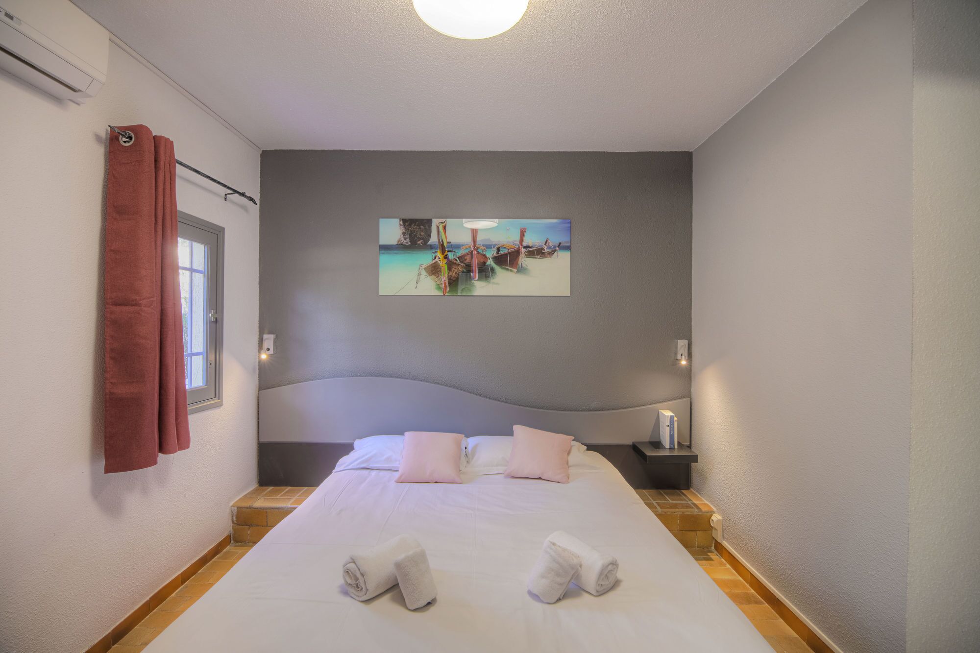Un dormitorio con cama, almohadas, un cuadro, una ventana con cortinas y una mesita de noche.