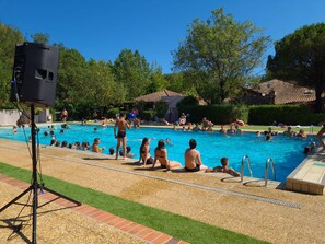 A swimming pool with people around it, a speaker on a tripod, and a grassy area with trees and buildings in the background. - Marina Hotel Club (Cogolin)